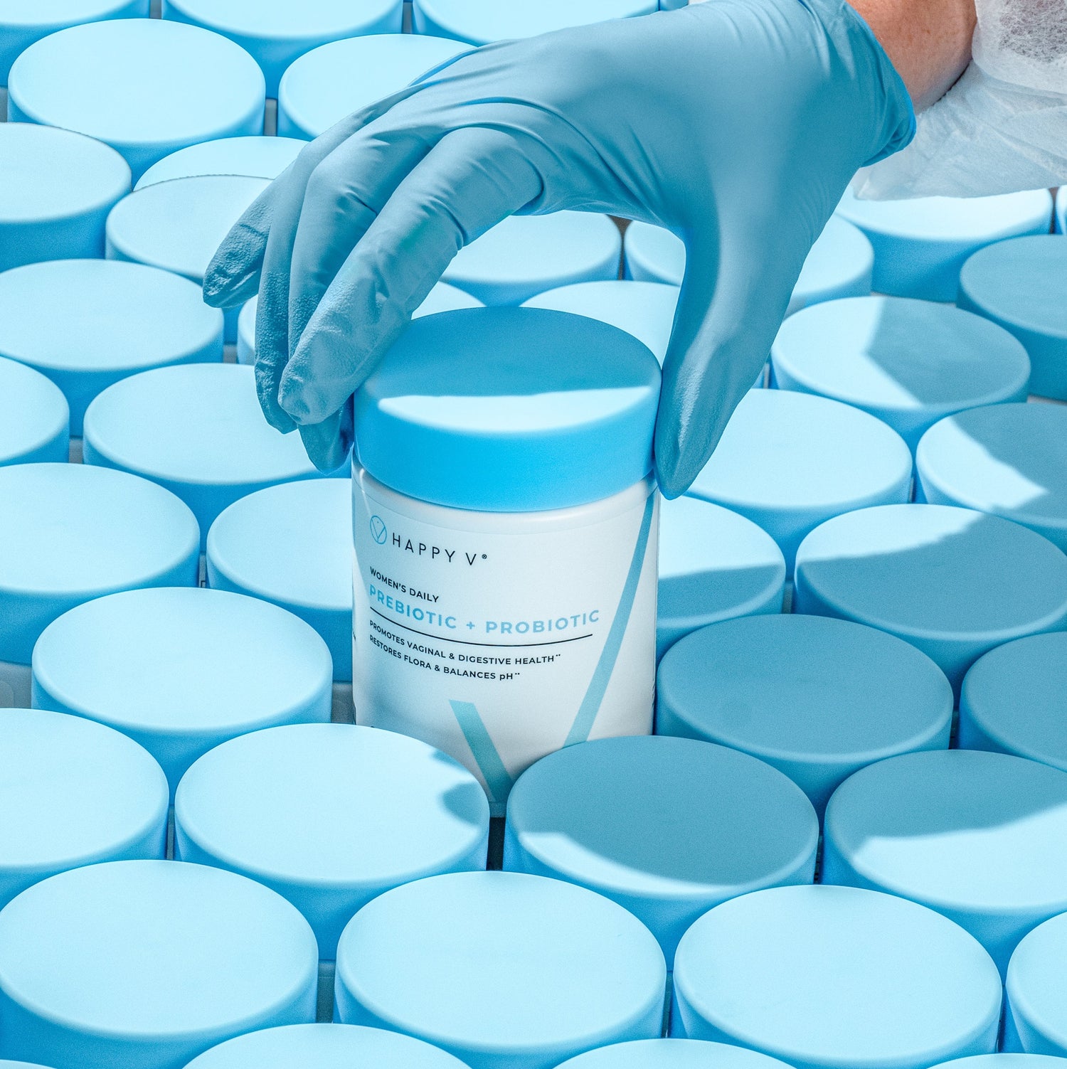 Hand placing Happy V probiotic bottle among scattered blue caps, highlighting clean packaging.